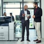 Zwei Männer in einem Büro diskutieren etwas auf einem Tablet neben einem Farbmultifunktionsdrucker der Xerox® AltaLink® C8200-Serie Zwei Männer in einem Büro diskutieren etwas auf einem Tablet neben einem Farbmultifunktionsdrucker der Xerox® AltaLink® C8200-Serie