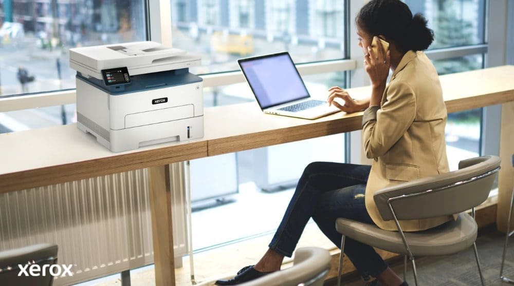 Xerox® Druck- und Scan-Erlebnis: Komfortabel und vernetzt Eine Person sitzt an einem hohen Tisch neben einem großen Fenster und telefoniert, während sie an ihrem Laptop arbeitet. Ein Multifunktionsdrucker Xerox B235DNI steht bequem auf dem Tisch daneben und suggeriert so eine einfache Bedienung in einem modernen Arbeitsbereich.