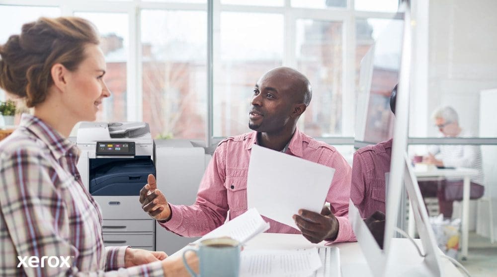 Xerox® Workplace Cloud und Suite: Zentralisiertes Flottenmanagement Eine Person überprüft Daten auf einem Laptop-Bildschirm mit einer digitalen Überlagerung verschiedener Gerätesymbole (Drucker, Mobilgeräte) und verbundener Linien, die das zentralisierte Flottenmanagement für Xerox Workplace Solutions symbolisieren.